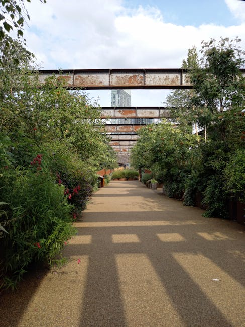 A pathway in a cultivated garden flanked by lush, well-maintained flowering shrubs and greenery on both sides, with a mix of blooming plants and trimmed foliage. The garden features a sandy-colored gr